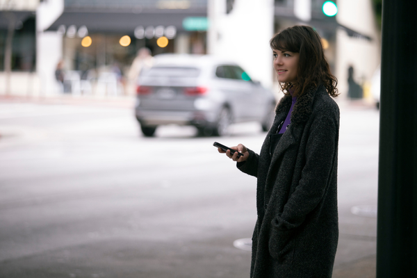 Woman with cellphone in hand, waiting on the side of a street with cars, appears to be waiting for a ride. 